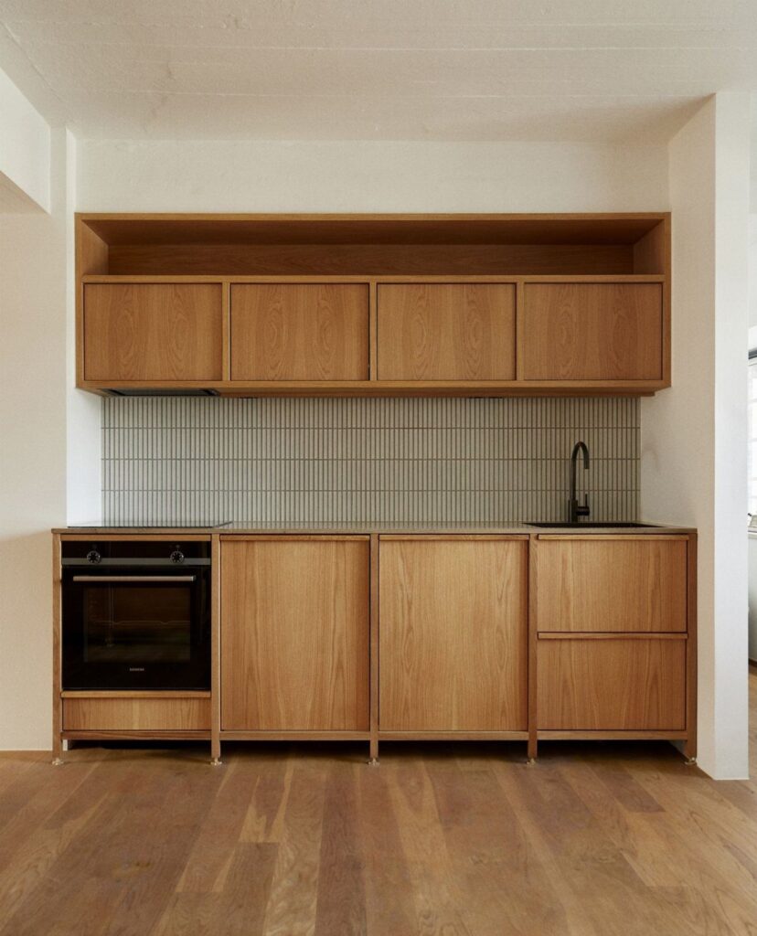 Minimalist kitchen with natural wood cabinets, wood flooring, and grey tile backsplash.