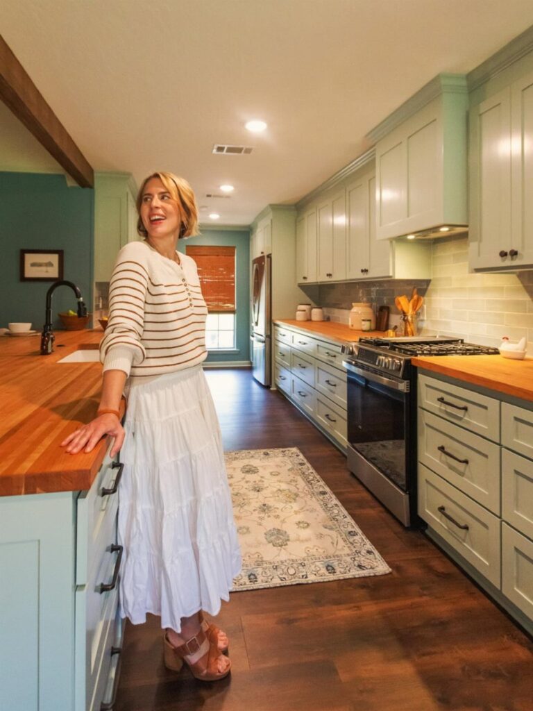 Woman standing in a kitchen with light green cabinets and butcher block countertops.